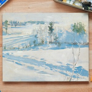 A winter landscape painting sits on a wooden table surrounded by paintbrushes and art supplies. The scene depicts snowy fields with trees, creating a serene and calm atmosphere.