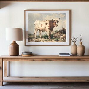 A wooden console table with a lamp, pottery, and books below a cow painting. Natural tones create a calm, rustic ambiance.