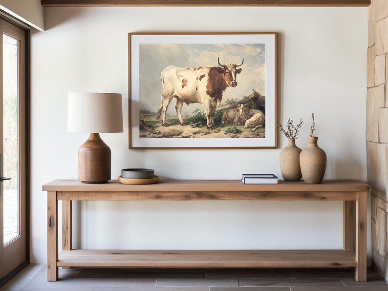 A wooden console table with a lamp, pottery, and books below a cow painting. Natural tones create a calm, rustic ambiance.