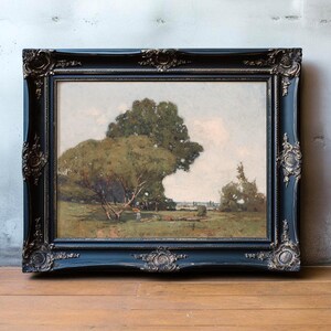 A landscape painting of a park scene with large, leafy trees and a person sitting on the grass, framed in an ornate black and silver frame, leaning against a textured, light-colored wall on a wooden floor.