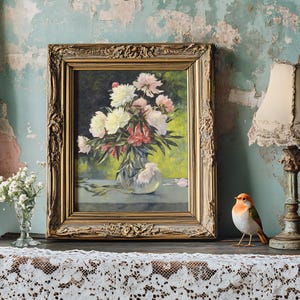 Framed floral painting on an antique table with lace cloth, beside a vintage lamp and bird figurine. The setting is rustic and charming.