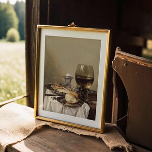 A framed still life painting rests on a rustic surface beside a chair. It depicts a wine glass, bread, and fruit on a table, evoking a serene, classical ambiance.