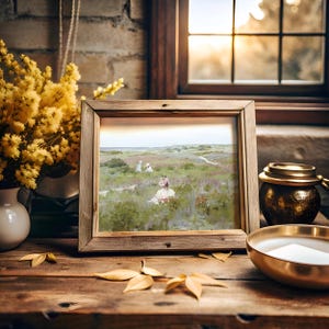 Framed painting of a serene landscape with figures in a field is on a rustic wooden table. Beside it are yellow flowers and a lit candle, creating a warm, cozy ambiance.