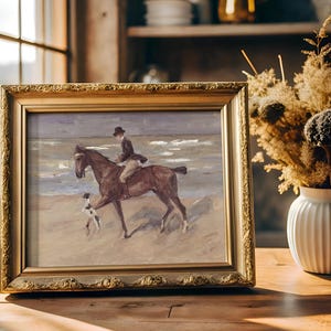 A framed painting of a person riding a horse on a beach with a dog, placed on a wooden table near a window. A vase with dried flowers is beside it.
