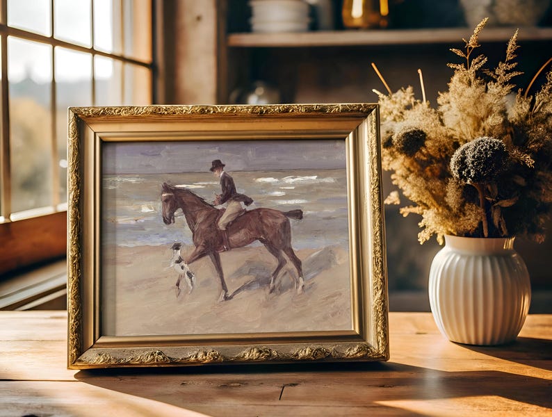 A framed painting of a person riding a horse on a beach with a dog, placed on a wooden table near a window. A vase with dried flowers is beside it.