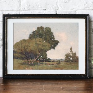 A framed landscape painting of a rural scene with large green trees, a person walking, and an open field under a soft sky, displayed on a wooden surface against a white brick wall. Next to the frame, there is a small potted cactus plant.