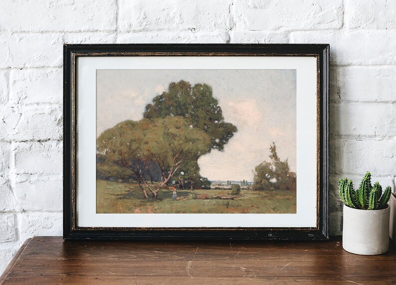 A framed landscape painting of a rural scene with large green trees, a person walking, and an open field under a soft sky, displayed on a wooden surface against a white brick wall. Next to the frame, there is a small potted cactus plant.