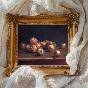 A framed still life painting of apples on a wooden table, surrounded by soft white fabric. The scene conveys a classic and serene tone.