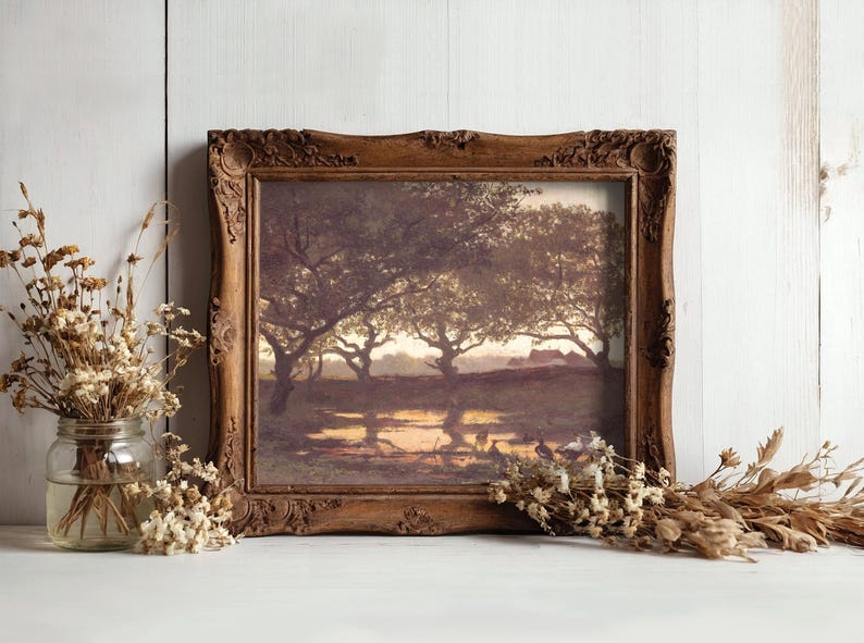 Framed landscape painting of trees at sunset, reflecting in a pond. Rustic wooden frame. Dried flowers in a jar and scattered on white surface.