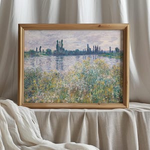 A framed impressionist painting of a river scene with trees and wildflowers in the foreground, displayed on a table covered with a light-colored cloth, with soft white curtains in the background.