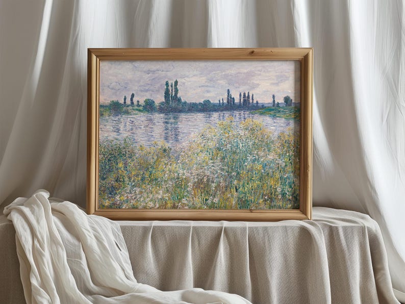 A framed impressionist painting of a river scene with trees and wildflowers in the foreground, displayed on a table covered with a light-colored cloth, with soft white curtains in the background.
