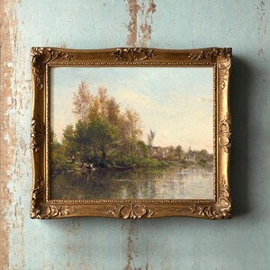 A framed landscape painting on a textured, weathered blue wall. The serene scene features a river, lush trees, and a quaint village, evoking calmness.
