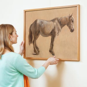 A woman in a light blue sweater carefully hangs a framed painting of a horse on a white wall. The painting shows a realistic brown horse on a beige background.