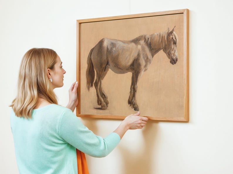 A woman in a light blue sweater carefully hangs a framed painting of a horse on a white wall. The painting shows a realistic brown horse on a beige background.