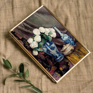 A framed painting of white roses in a blue vase, placed on a textured beige fabric. A small green branch lies beside it. The words "PRINTS FARM" appear above.