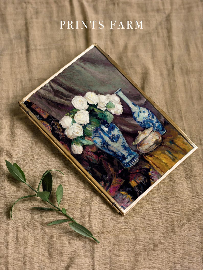 A framed painting of white roses in a blue vase, placed on a textured beige fabric. A small green branch lies beside it. The words "PRINTS FARM" appear above.