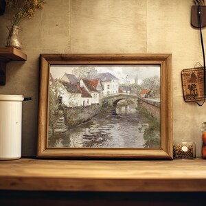 A framed painting of a scenic village by a bridge over a river sits on a kitchen shelf, surrounded by jars, a vase, and rustic decor, evoking a cozy, nostalgic vibe.