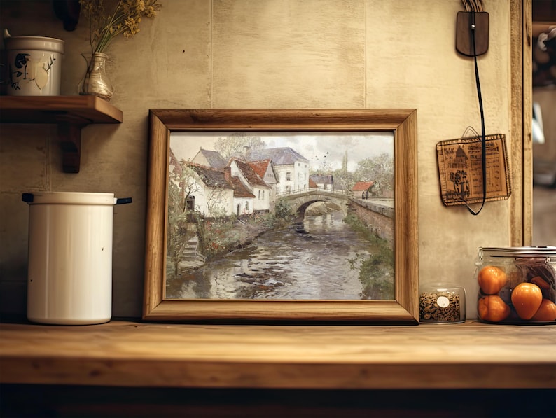 A framed painting of a scenic village by a bridge over a river sits on a kitchen shelf, surrounded by jars, a vase, and rustic decor, evoking a cozy, nostalgic vibe.