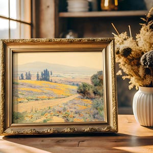 A framed landscape painting on a sunlit wooden table near a window, depicting a serene countryside with fields and trees. A vase of dried flowers is nearby.