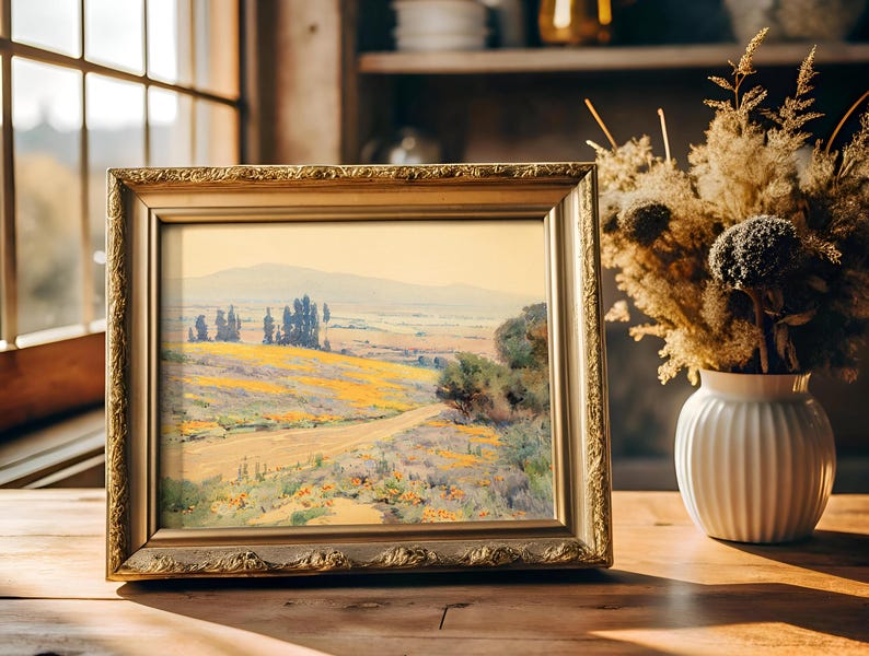 A framed landscape painting on a sunlit wooden table near a window, depicting a serene countryside with fields and trees. A vase of dried flowers is nearby.