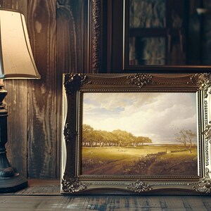 A framed landscape painting depicting a serene countryside with trees and a vast sky rests on a wooden table beside an elegant table lamp.
