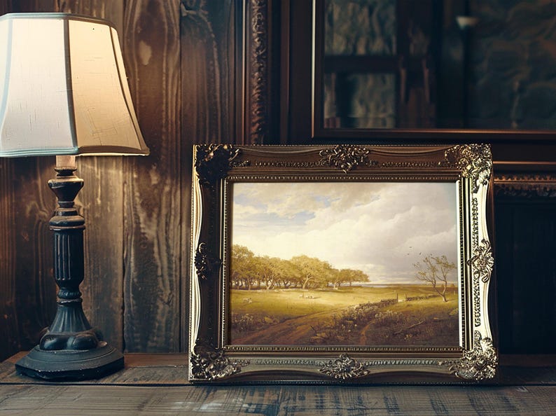 A framed landscape painting depicting a serene countryside with trees and a vast sky rests on a wooden table beside an elegant table lamp.