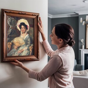 A woman carefully hangs a framed painting of an elegant lady with a fan on a wall in a cozy living room. The setting is warm and inviting.