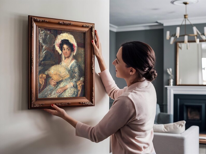 A woman carefully hangs a framed painting of an elegant lady with a fan on a wall in a cozy living room. The setting is warm and inviting.