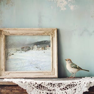 A rustic painting of a snowy village in a vintage frame sits on a wooden shelf. Beside it, a small bird is perched, with lace fabric underneath, creating a serene, cozy scene.