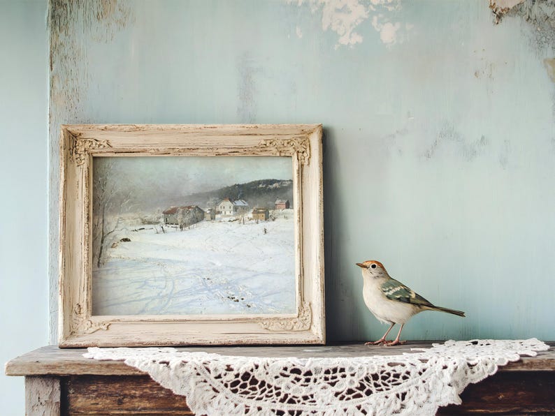 A rustic painting of a snowy village in a vintage frame sits on a wooden shelf. Beside it, a small bird is perched, with lace fabric underneath, creating a serene, cozy scene.