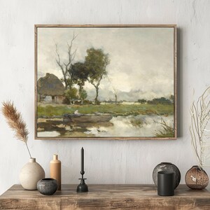 Framed landscape painting of a serene rural scene with a house and trees by a river. Rustic decor on wooden table below evokes calmness.