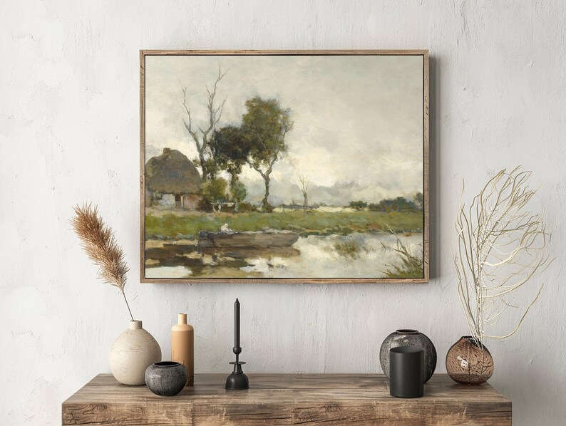 Framed landscape painting of a serene rural scene with a house and trees by a river. Rustic decor on wooden table below evokes calmness.