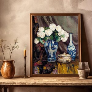 A framed painting of white flowers in a blue vase is displayed on a rustic wooden table. It is flanked by a candle, pottery, and dried flowers.