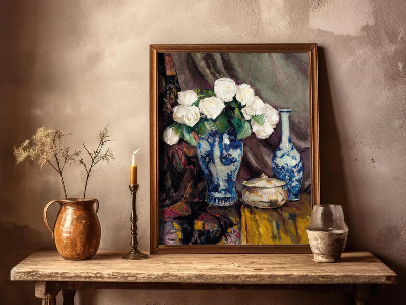 A framed painting of white flowers in a blue vase is displayed on a rustic wooden table. It is flanked by a candle, pottery, and dried flowers.