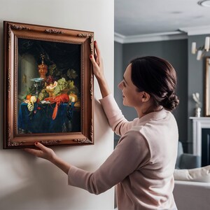 A woman in a light sweater hangs a framed still-life painting with fruits and a lobster on a beige wall in a modern living room, conveying elegance and warmth.