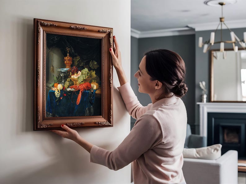 A woman in a light sweater hangs a framed still-life painting with fruits and a lobster on a beige wall in a modern living room, conveying elegance and warmth.