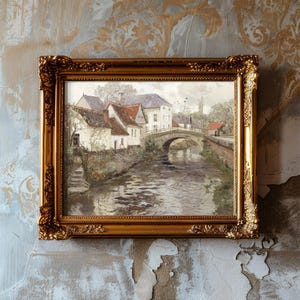 A framed landscape painting with a gold ornate border hangs on a textured, aged wall. The artwork depicts a serene village scene with white cottages, a stone bridge, and a gently flowing river, evoking a sense of nostalgia and tranquility.