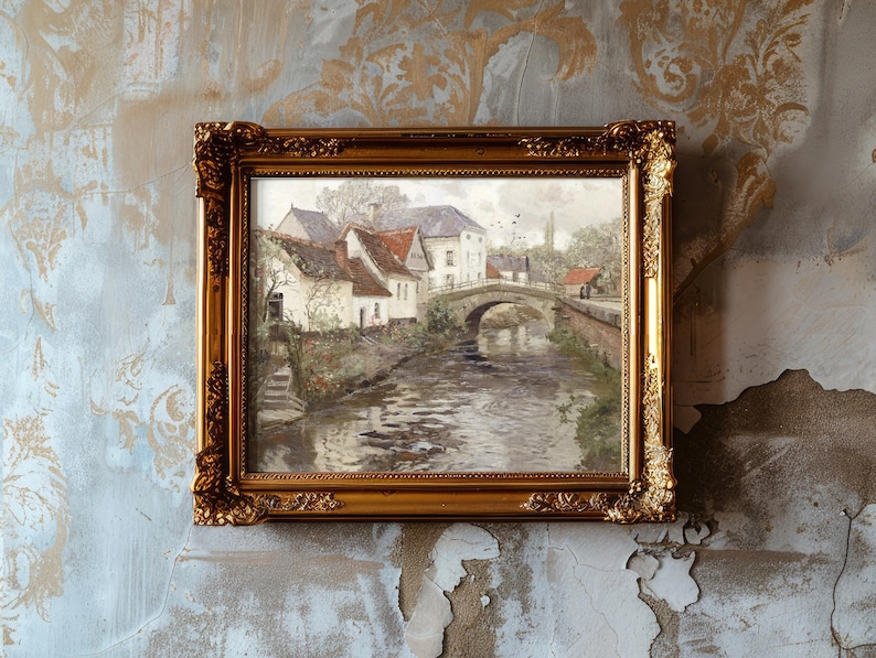 A framed landscape painting with a gold ornate border hangs on a textured, aged wall. The artwork depicts a serene village scene with white cottages, a stone bridge, and a gently flowing river, evoking a sense of nostalgia and tranquility.
