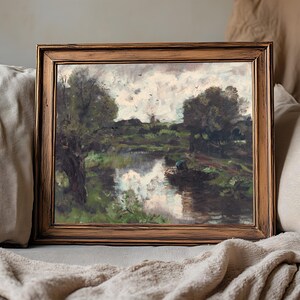 A framed landscape painting rests on a beige couch. It depicts a serene river scene with trees, cloudy sky, and reflections on the water, evoking tranquility.