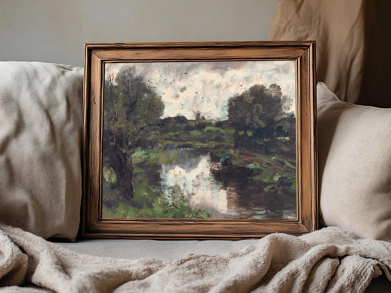 A framed landscape painting rests on a beige couch. It depicts a serene river scene with trees, cloudy sky, and reflections on the water, evoking tranquility.