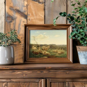 Framed pastoral painting of sheep set on a rustic wooden shelf, flanked by a potted succulent and leafy plant, creating a serene, natural ambiance.