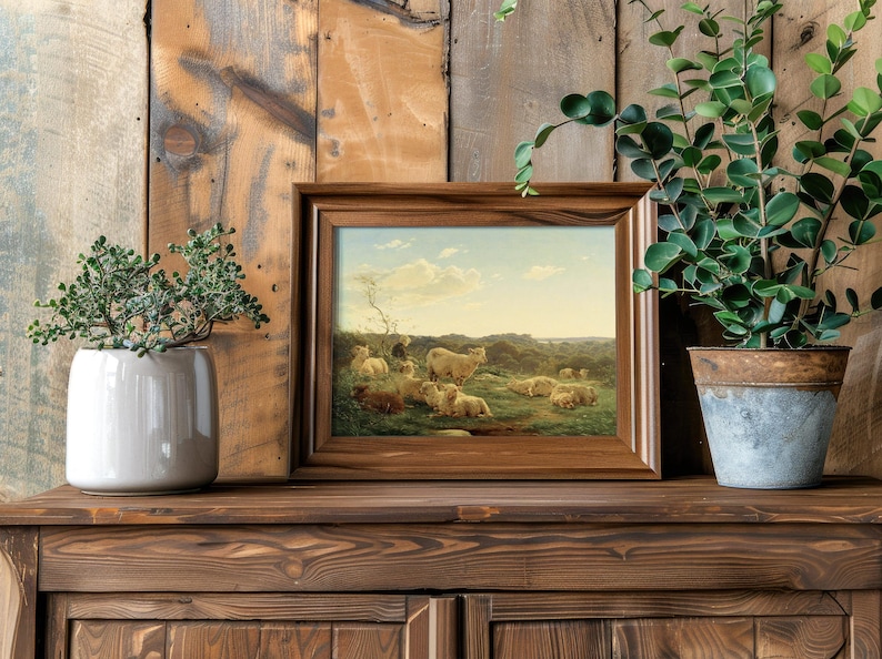 Framed pastoral painting of sheep set on a rustic wooden shelf, flanked by a potted succulent and leafy plant, creating a serene, natural ambiance.