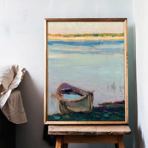 A framed painting of a peaceful lakeside scene with a small boat on the shore, resting on a wooden easel in a softly lit room with a light blue wall.