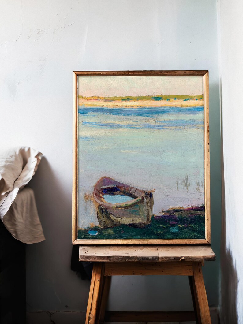 A framed painting of a peaceful lakeside scene with a small boat on the shore, resting on a wooden easel in a softly lit room with a light blue wall.