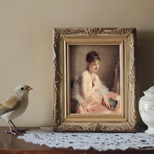 A delicate bird stands on a lace-covered table next to a vintage framed painting of a seated woman with a gentle expression, alongside an ornate ceramic jar.