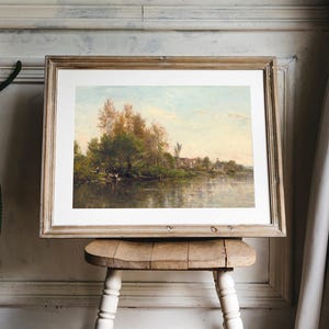 A framed landscape painting depicting a river scene with trees and a small village in the background. The artwork is displayed on a small, rustic wooden stool. The frame is a light brown color, and the painting has a soft, natural color palette.