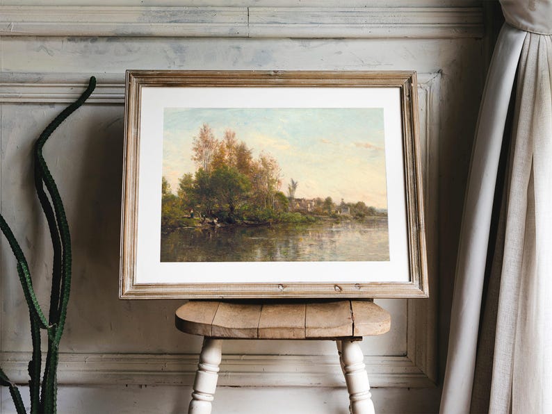A framed landscape painting depicting a river scene with trees and a small village in the background. The artwork is displayed on a small, rustic wooden stool. The frame is a light brown color, and the painting has a soft, natural color palette.