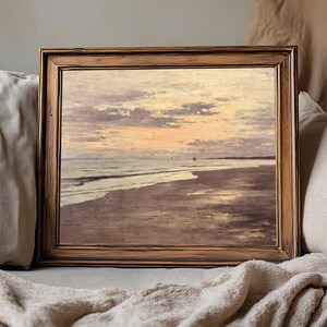Framed beach painting with a serene sunset, brown wooden frame resting on a cozy beige couch, conveying a calm and tranquil ambiance.