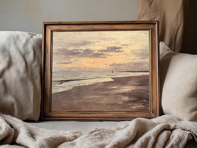 Framed beach painting with a serene sunset, brown wooden frame resting on a cozy beige couch, conveying a calm and tranquil ambiance.