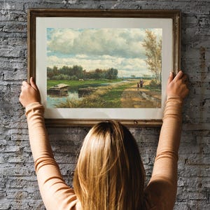 A person hangs a framed landscape painting on a brick wall. The painting depicts a rural scene with a river, trees, and a cloudy sky.